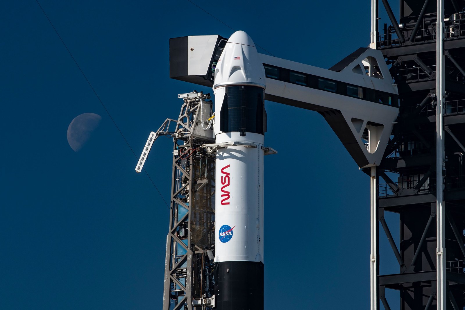 Crew Dragon atop Falcon 9 at the pad, with the crew access arm and the moon behind